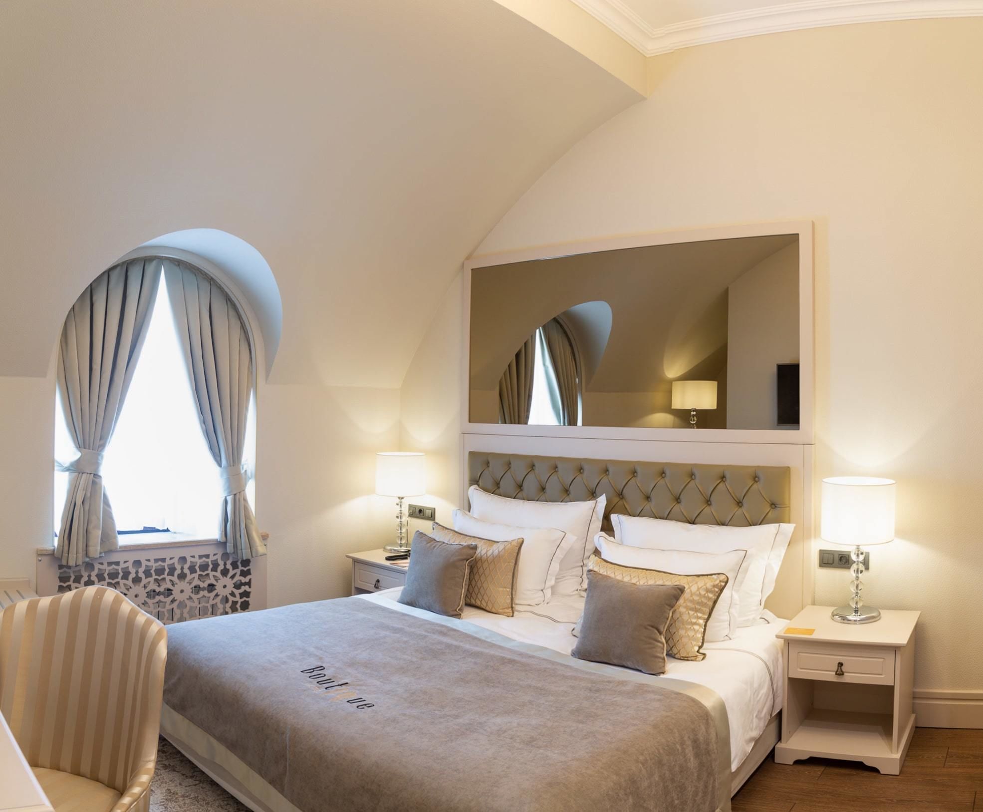 A bedroom with a large bed, two bedside tables, a chair, a mirror, and a window with curtains.