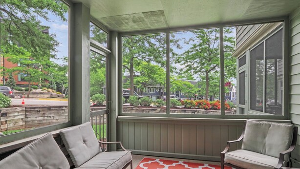 A porch with a gray sofa and a chair, a red patterned rug, and a view of a garden and buildings. - Pointes North Inn 109 | Downtown Charlevoix | Indoor pool (Charlevoix)