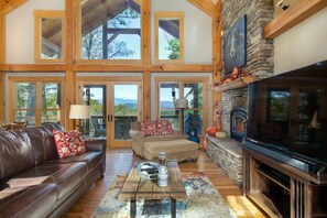 A spacious living room with a stone fireplace, leather sofas, and a large TV. - Timber Hideaway | Pet-Friendly Timber Frame Home with Hot Tub & Views! (Old Fort)