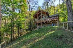 A two-story log cabin with a balcony, surrounded by a forested hillside. - Timber Hideaway | Pet-Friendly Timber Frame Home with Hot Tub & Views! (Old Fort)
