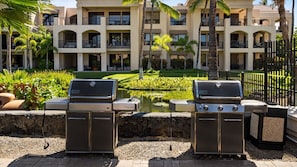 Two barbecue grills in a courtyard with a pool, palm trees, and a multi-story building in the background. - 🏖️Ground Floor | In-Unit Central AC | Concierge Included (Waikoloa)