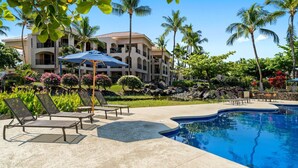 A resort pool area with lounge chairs and umbrellas, surrounded by palm trees and a large building in the background. - 🏖️Hidden Garden Feel | Large Lanai + A/C | Concierge Included (Waikoloa)