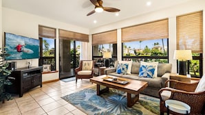 A living room with a television, sofa, coffee table, and a view of palm trees through large windows. - 🏖️Hidden Garden Feel | Large Lanai + A/C | Concierge Included (Waikoloa)