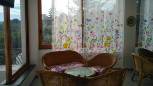Et rum med et glasbord omgivet af fletstole, et blomstret gardin og udsigt til grøn bevoksning udenfor.
