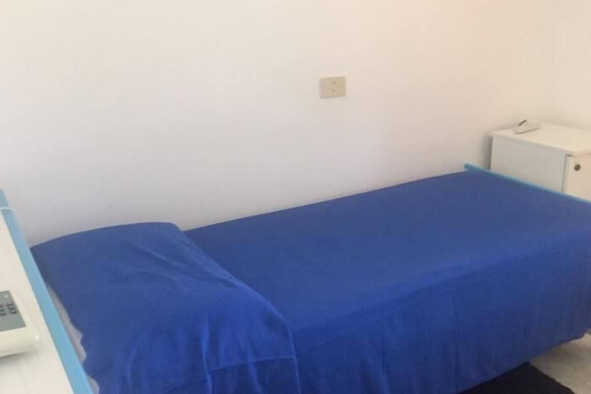 Une chambre avec un lit simple, une couverture bleue, une petite table de chevet et un interrupteur fixé au mur.