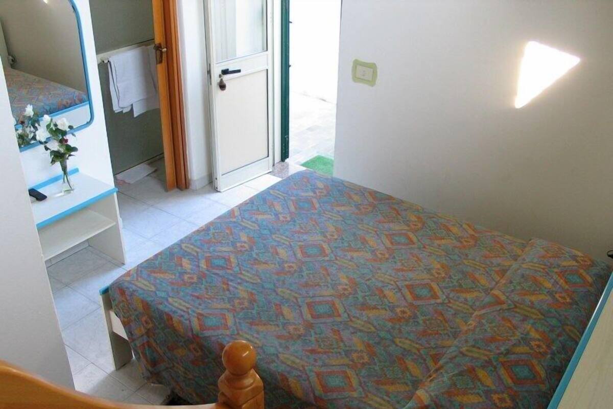 Une petite pièce simple, avec un lit, une chaise en bois, une petite table sur laquelle est posé un vase de fleurs, et une porte donnant sur un espace extérieur.