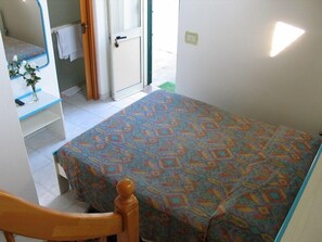 A small, simple room with a bed, a wooden chair, a small table with a vase of flowers, and a door leading to an outdoor area. - Villa Petrusa (Lipari)
