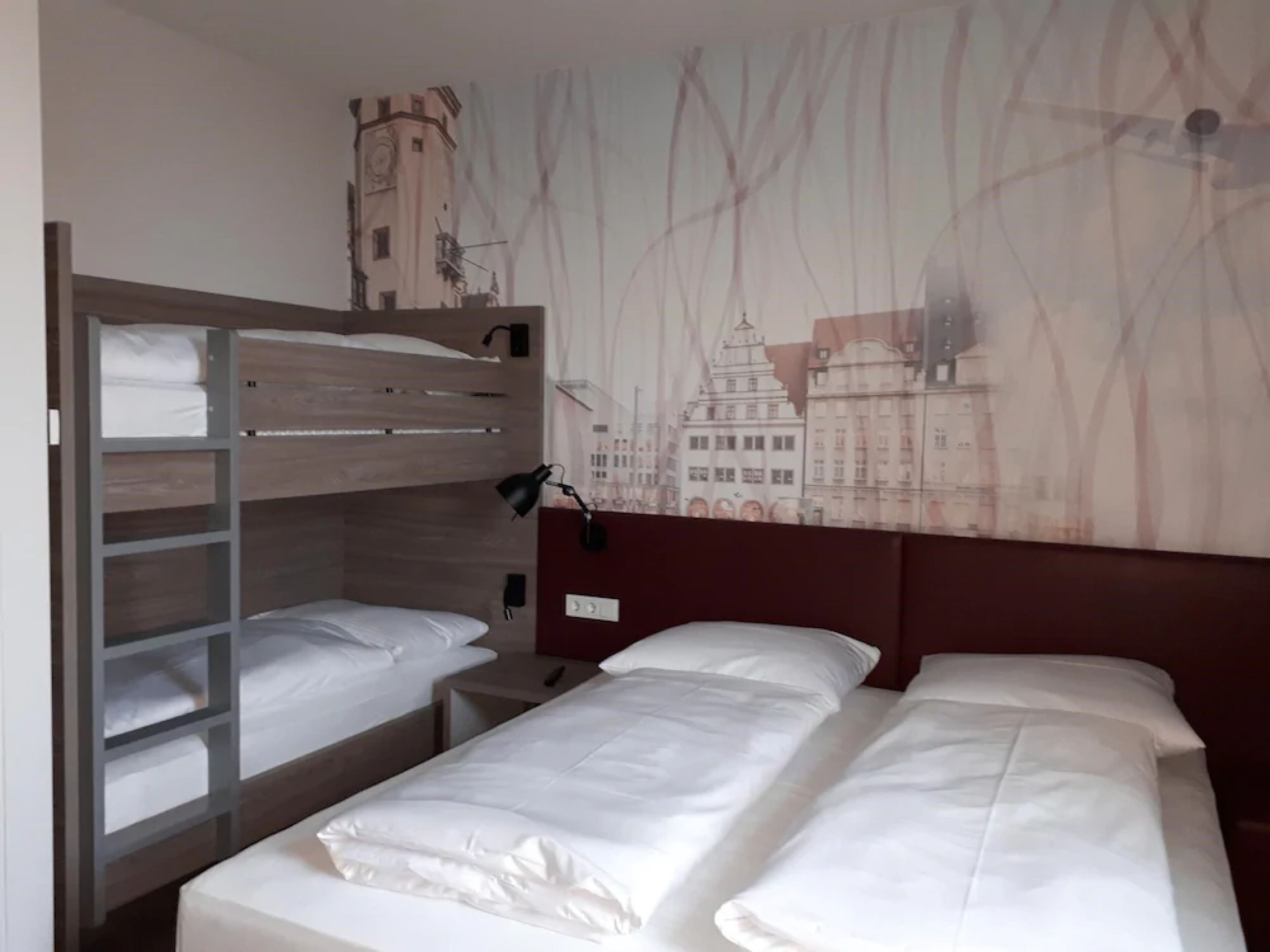 Et hotelværelse med køjeseng, enkeltseng, et natbord, en lampe og en væg med et bylandskabsmaleri.