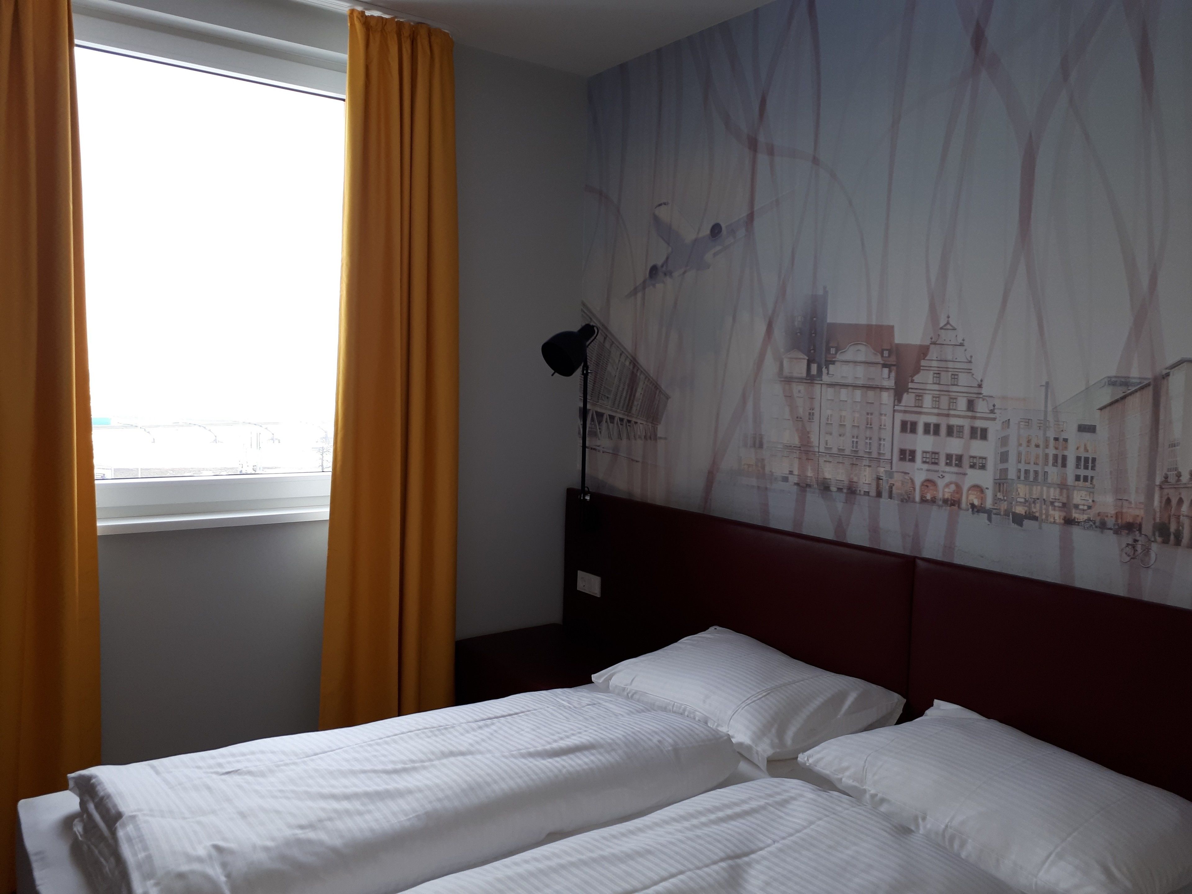 A hotel room with a bed, a window with yellow curtains, and a cityscape mural on the wall.