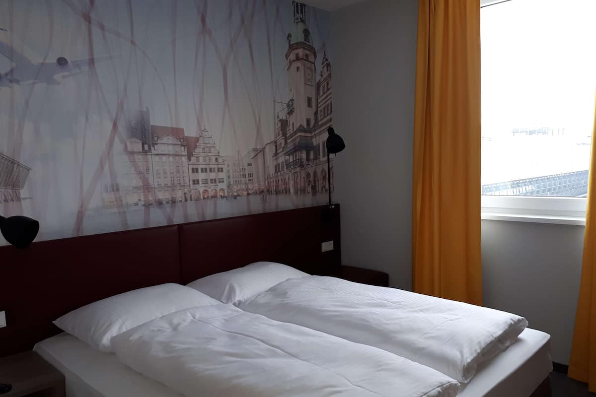 Ein Hotelzimmer mit Bett, Nachttisch, Lampe, einem Fenster mit Vorhängen und einem Wandbild einer Stadtansicht.