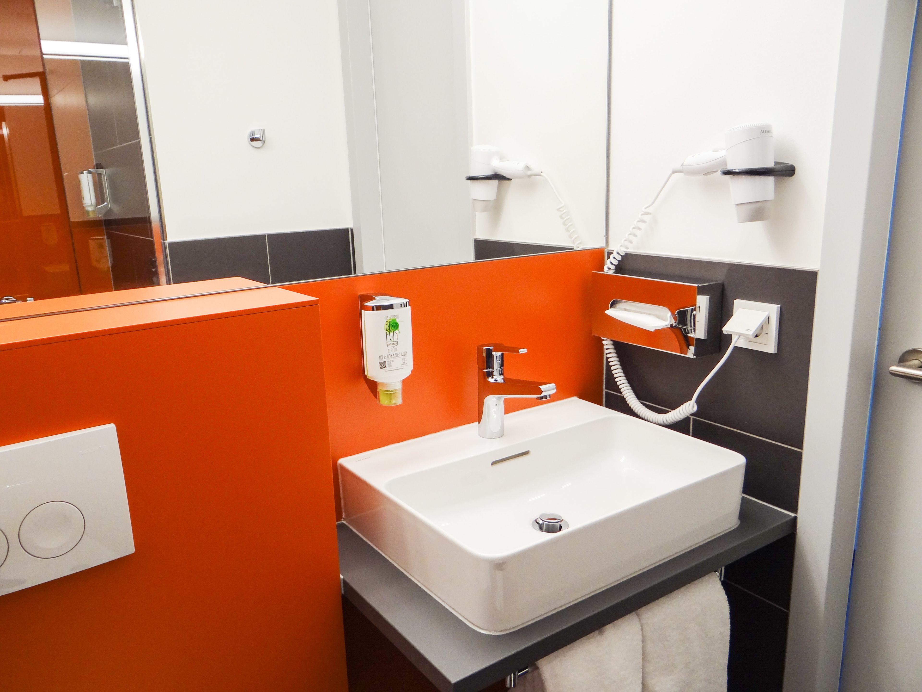 A modern bathroom with a white sink, orange accents, and a mirror.