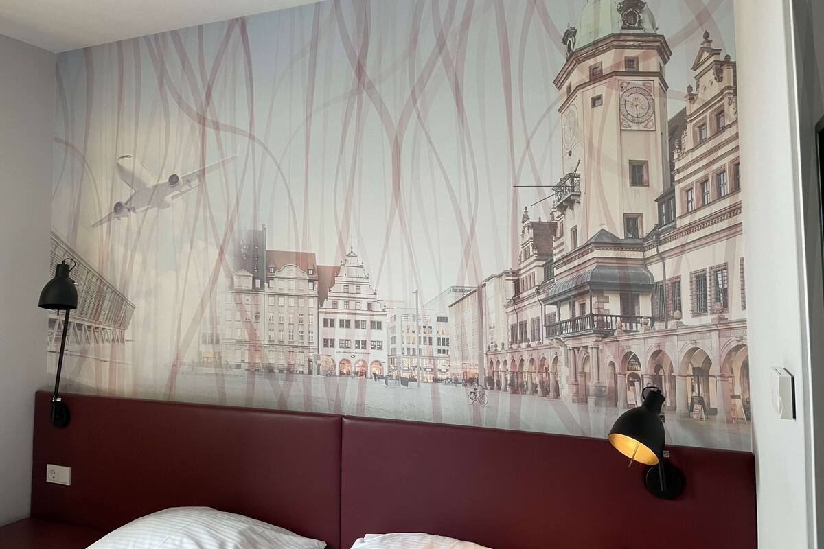 Ein Hotelzimmer mit einer großen Wandmalerei einer Stadtansicht, einem roten, gepolsterten Kopfteil und einer schwarzen Lampe auf dem Nachttisch.