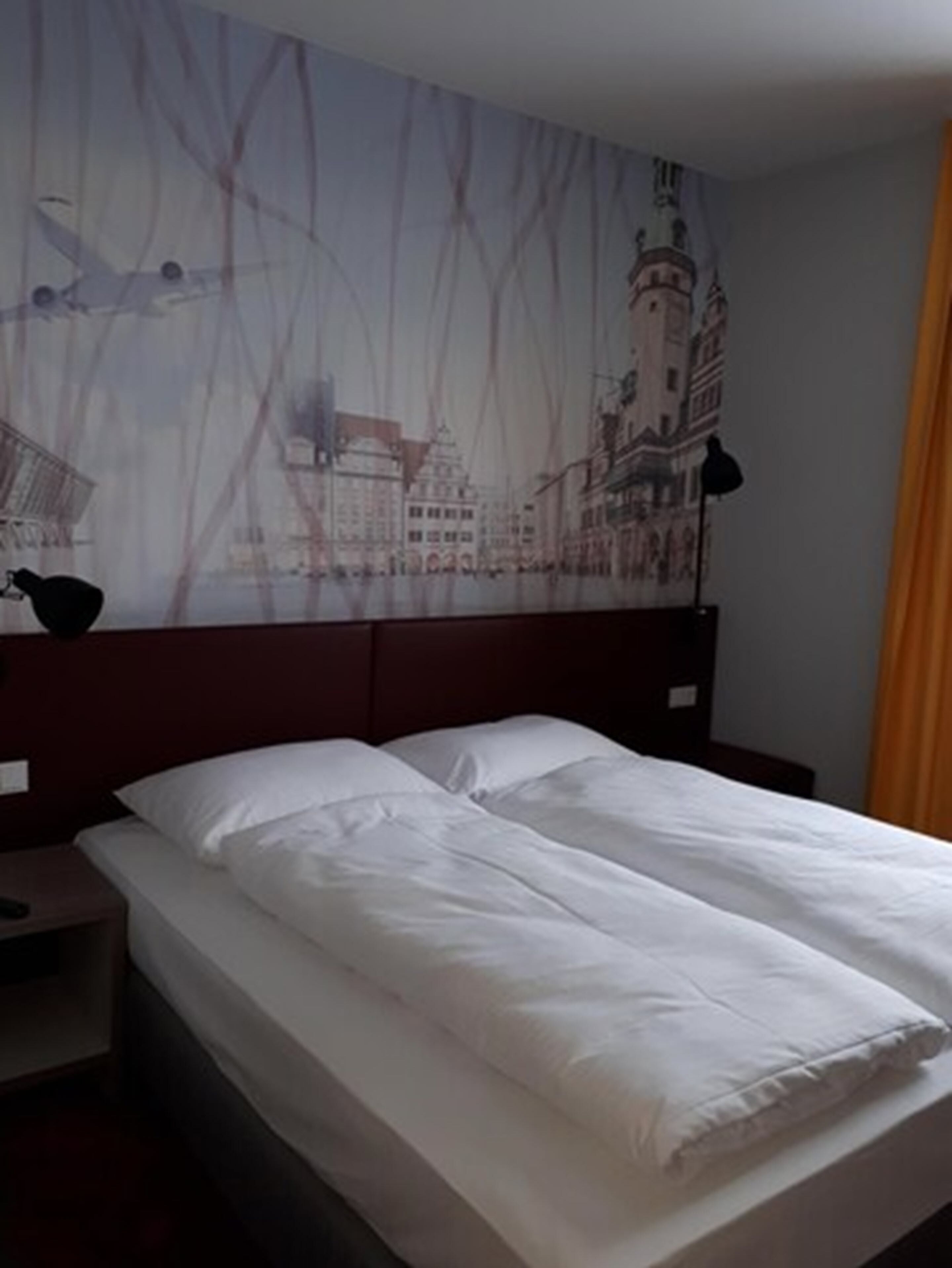 Ein Hotelzimmer mit einem Bett, einem Kopfteil, einem Nachttisch und einer Wand mit Stadtansicht-Mural.