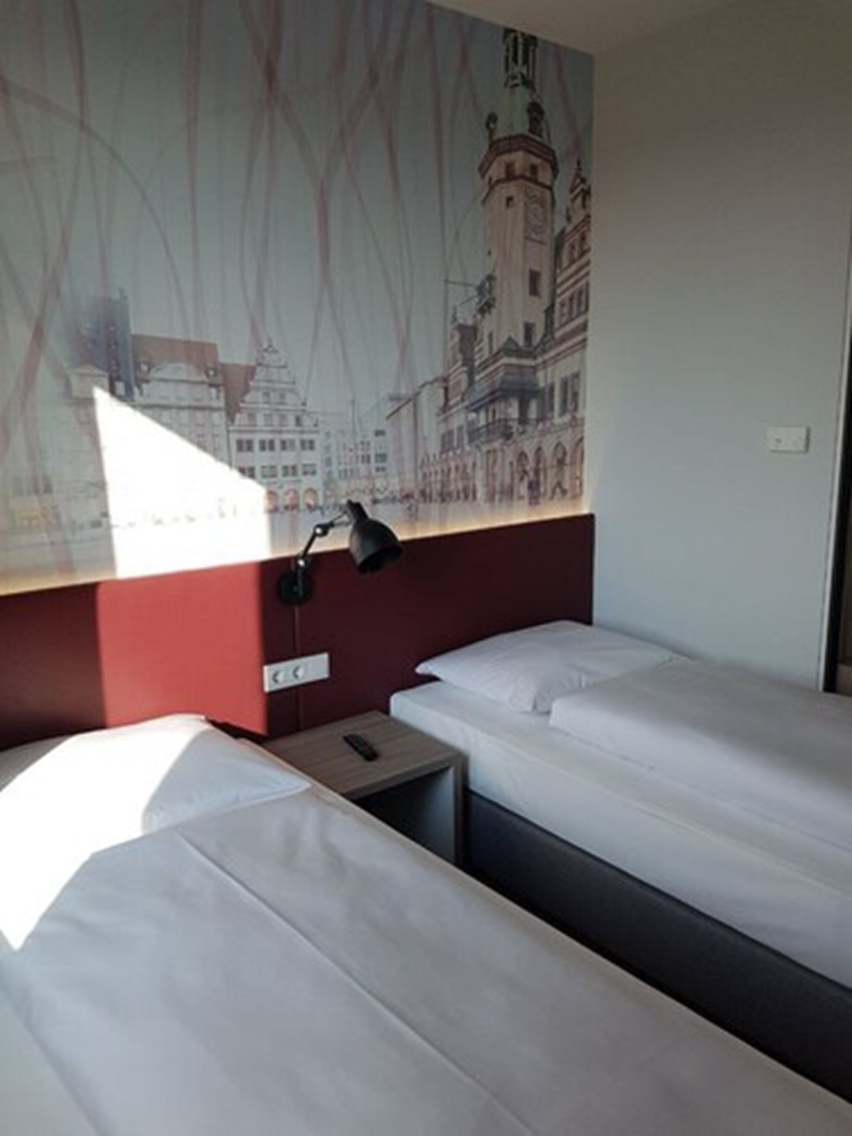 A hotel room with two single beds, a nightstand with a lamp, and a wall mural of a cityscape.