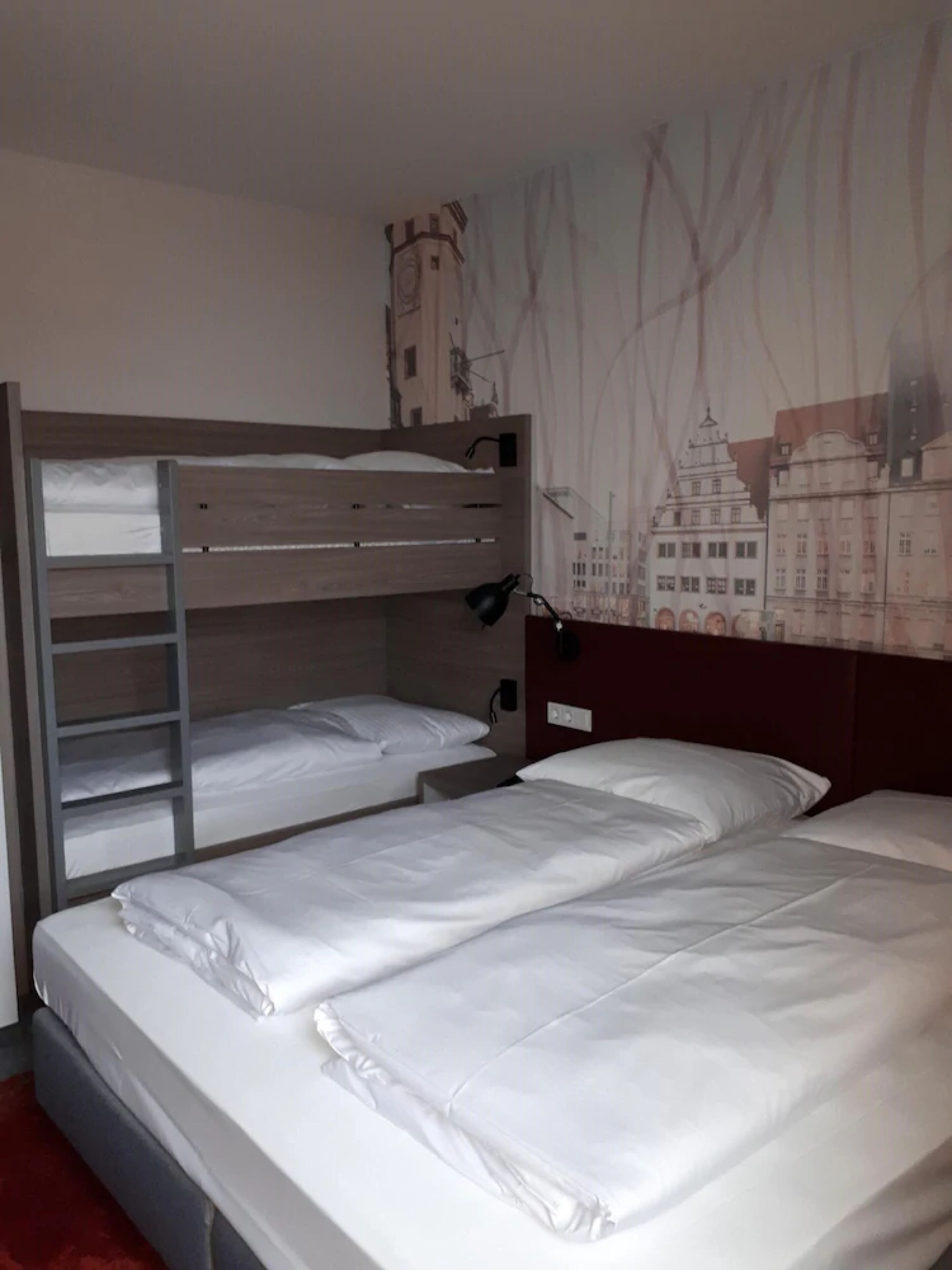 A hotel room with two single beds, a bunk bed, a red carpet, and a cityscape mural on the wall.