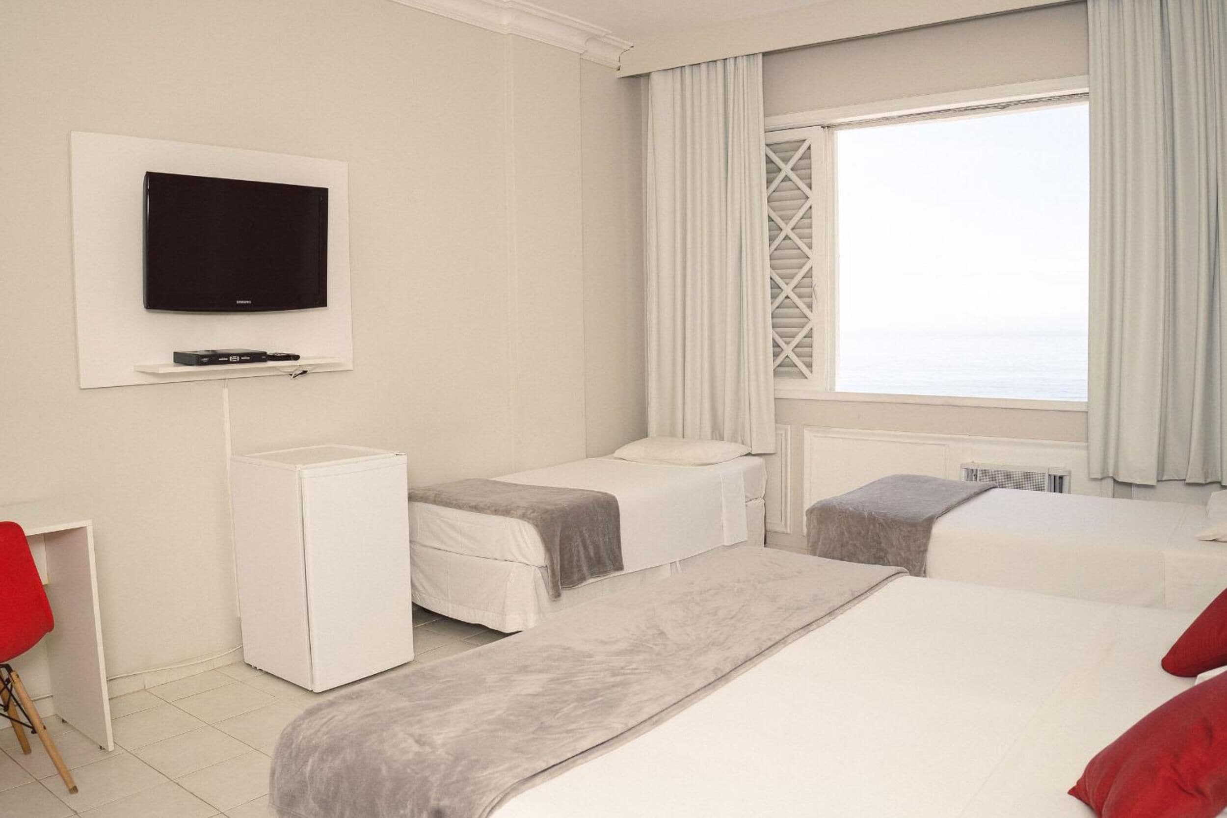 A hotel room with two beds, a TV mounted on the wall, a small white cabinet, a red chair, and a window with curtains.