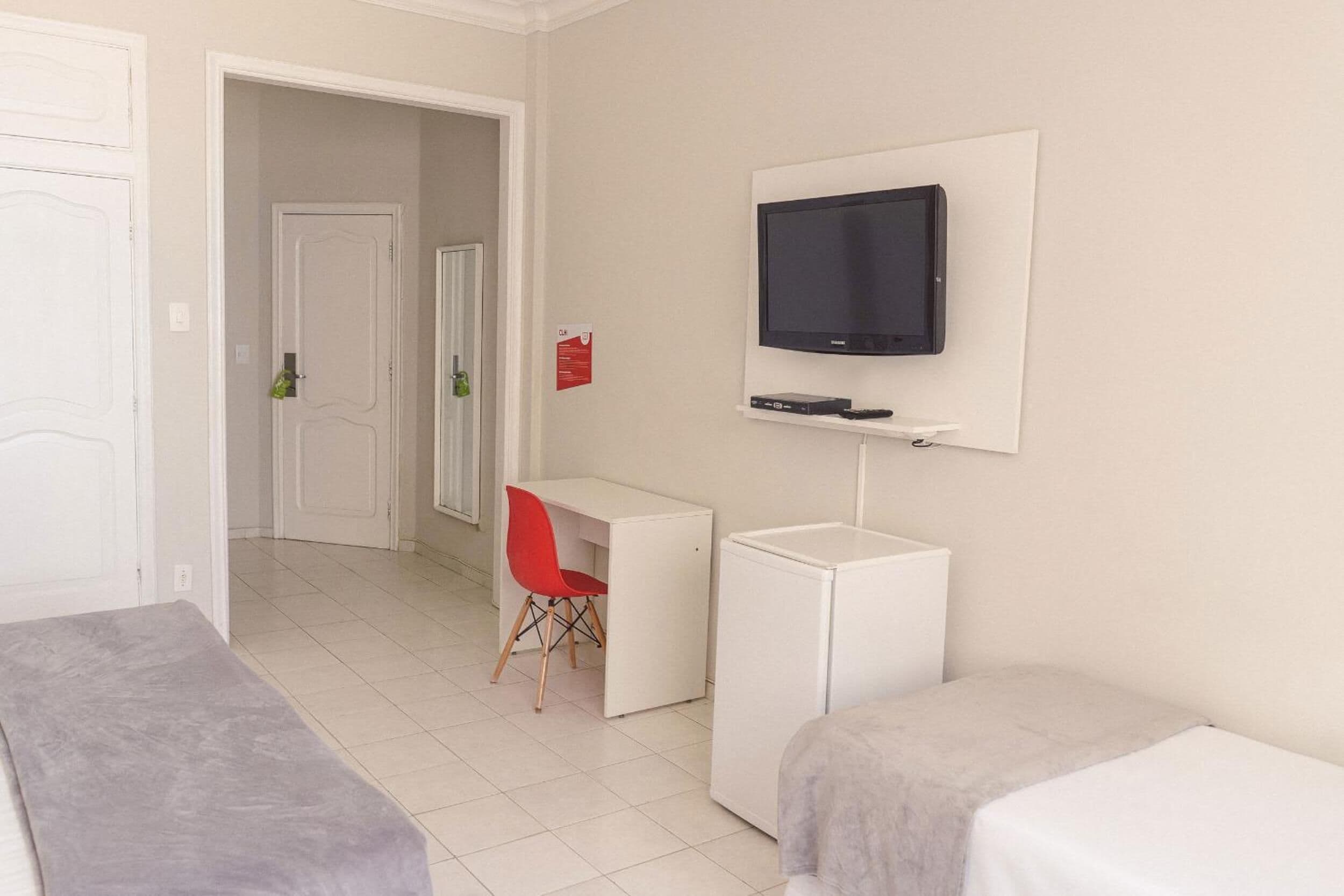 Quarto de hotel com cama, escrivaninha, cadeira, televisão e um pequeno frigorífico.