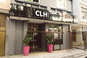 Entrada de un hotel de diseño moderno, con un llamativo letrero 'CLH Suites' sobre la puerta. Hay dos grandes macetas con plantas frente a la entrada, y un camino de acceso con puerta reja es visible a la derecha.