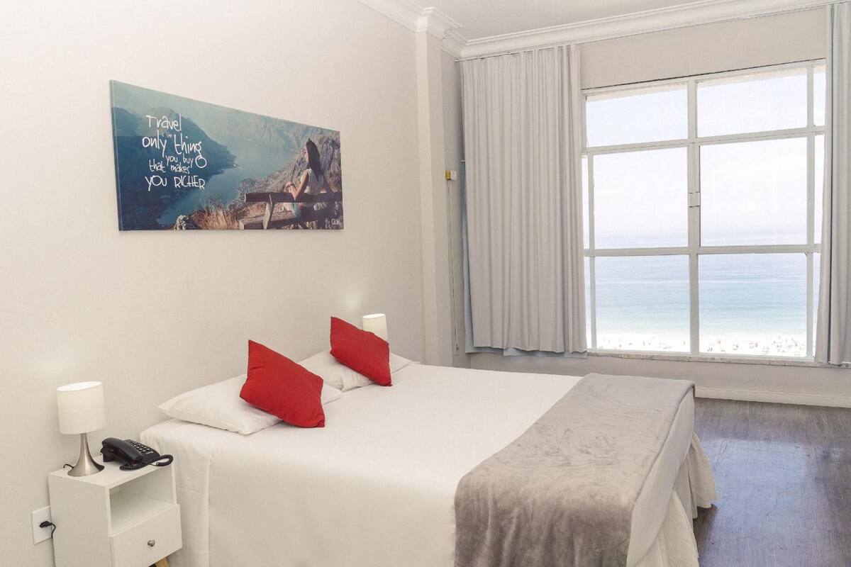 A hotel room with a bed, a nightstand with a lamp and phone, a painting on the wall, and a large window with a view of the sea.