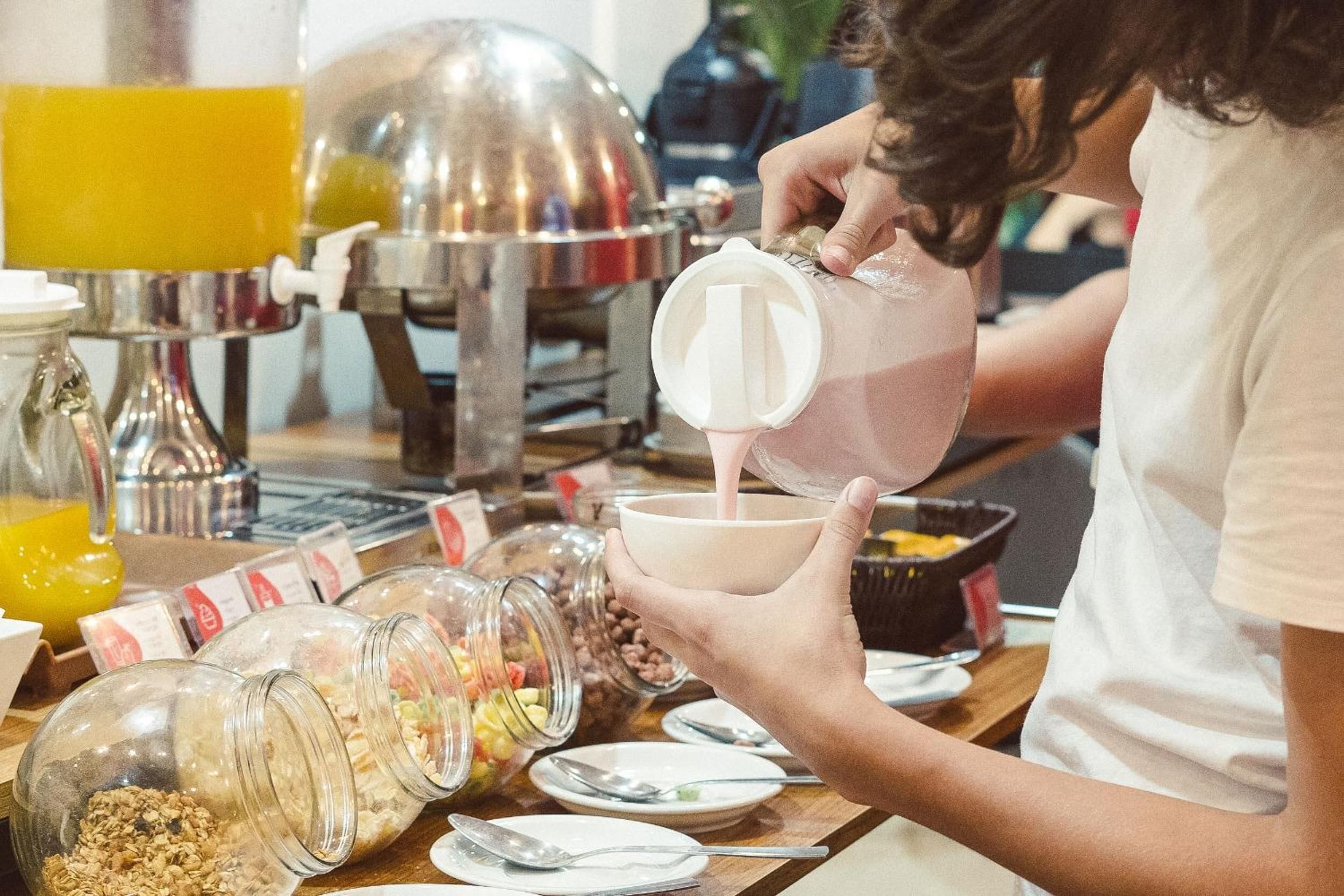 A breakfast setting with a person pouring a beverage into a bowl, surrounded by various food items and kitchen appliances.