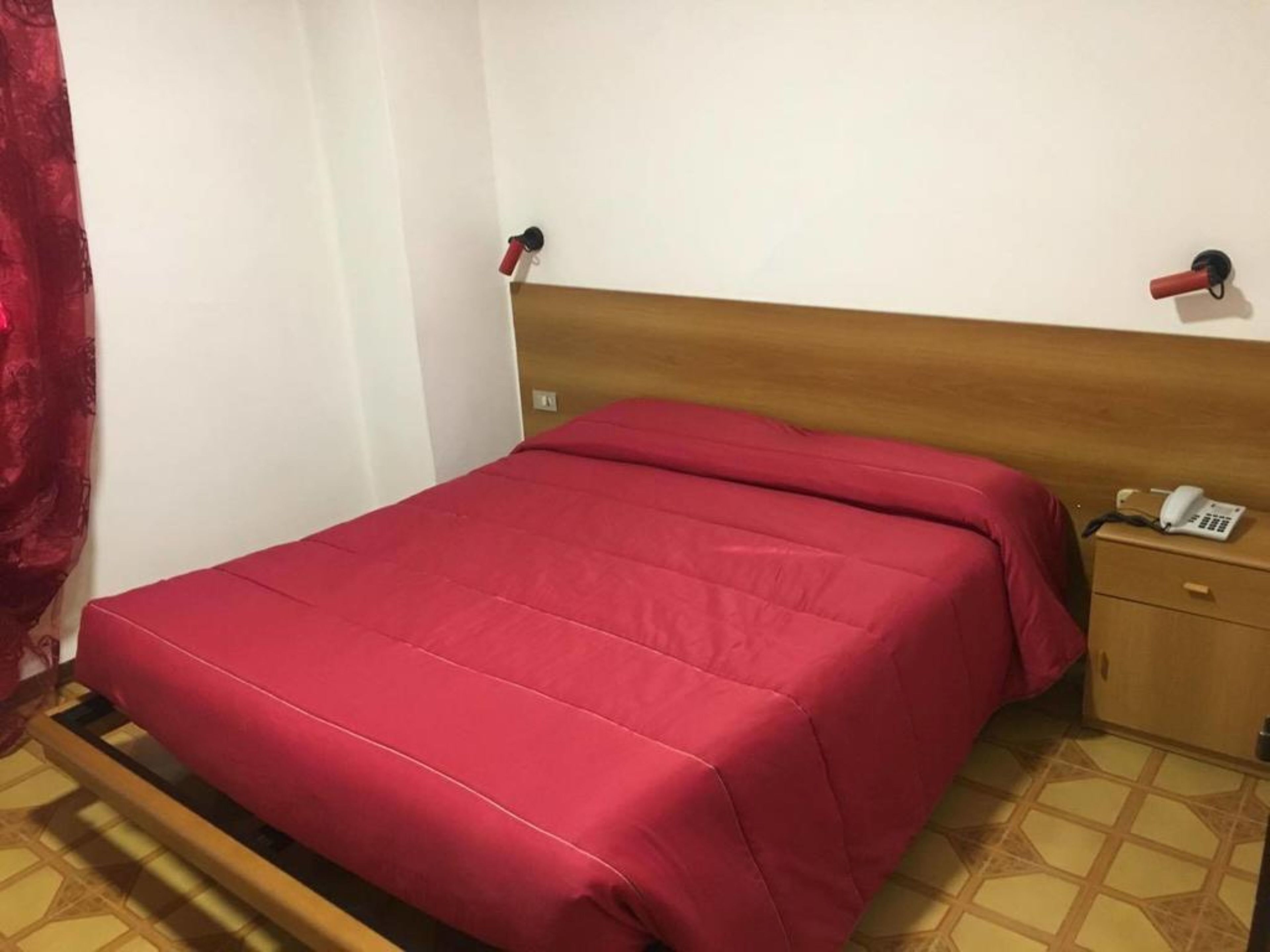A single-bed room with a wooden headboard, a red bedspread, a bedside table with a telephone, and a red curtain on the left.