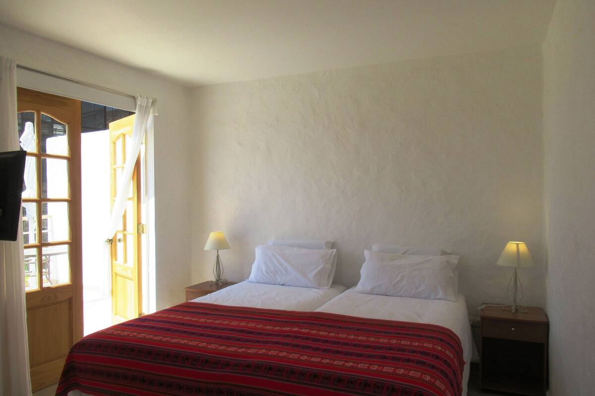 Un dormitorio con una cama, dos lámparas de noche, un ventanal con cortinas y una puerta de madera.