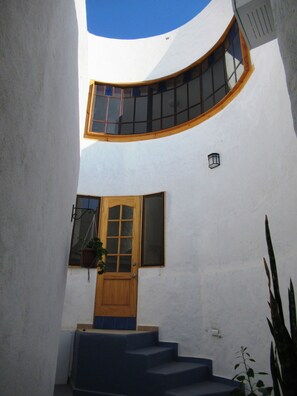 Una escalera curva que conduce a una puerta de madera con un ventanal encima, rodeada de paredes blancas y un cielo azul visible a través de la curva.