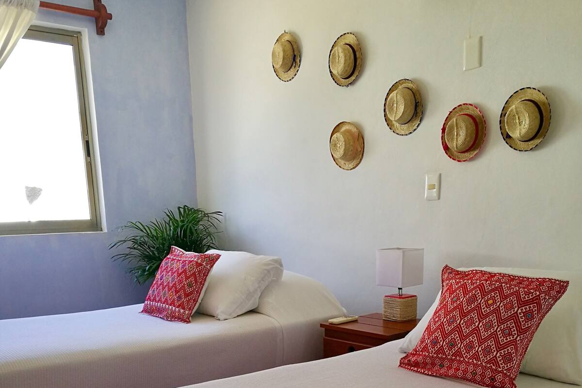 Ein Hotelzimmer mit zwei Betten, jeweils mit weißer Bettwäsche und roten, gemusterten Kissen. Sechs Hüte hängen an der Wand, ein Fenster mit Vorhängen und ein Nachttisch mit Lampe und Fernbedienung.
