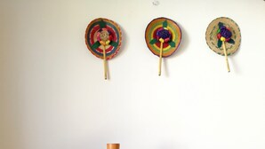 A round wooden table with a ceramic vase in the center, flanked by two woven chairs. Above the table, three colorful woven wall hangings are displayed.