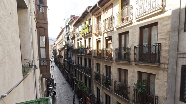 Una calle estrecha flanqueada por edificios de varios pisos, cada uno con balcones y plantas en macetas.