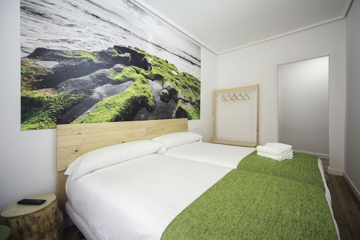 Un dormitorio con cama, cabecera de madera, manta verde, una obra de arte colgada en la pared que representa un paisaje rocoso, y una mesita de noche de madera con un control remoto.