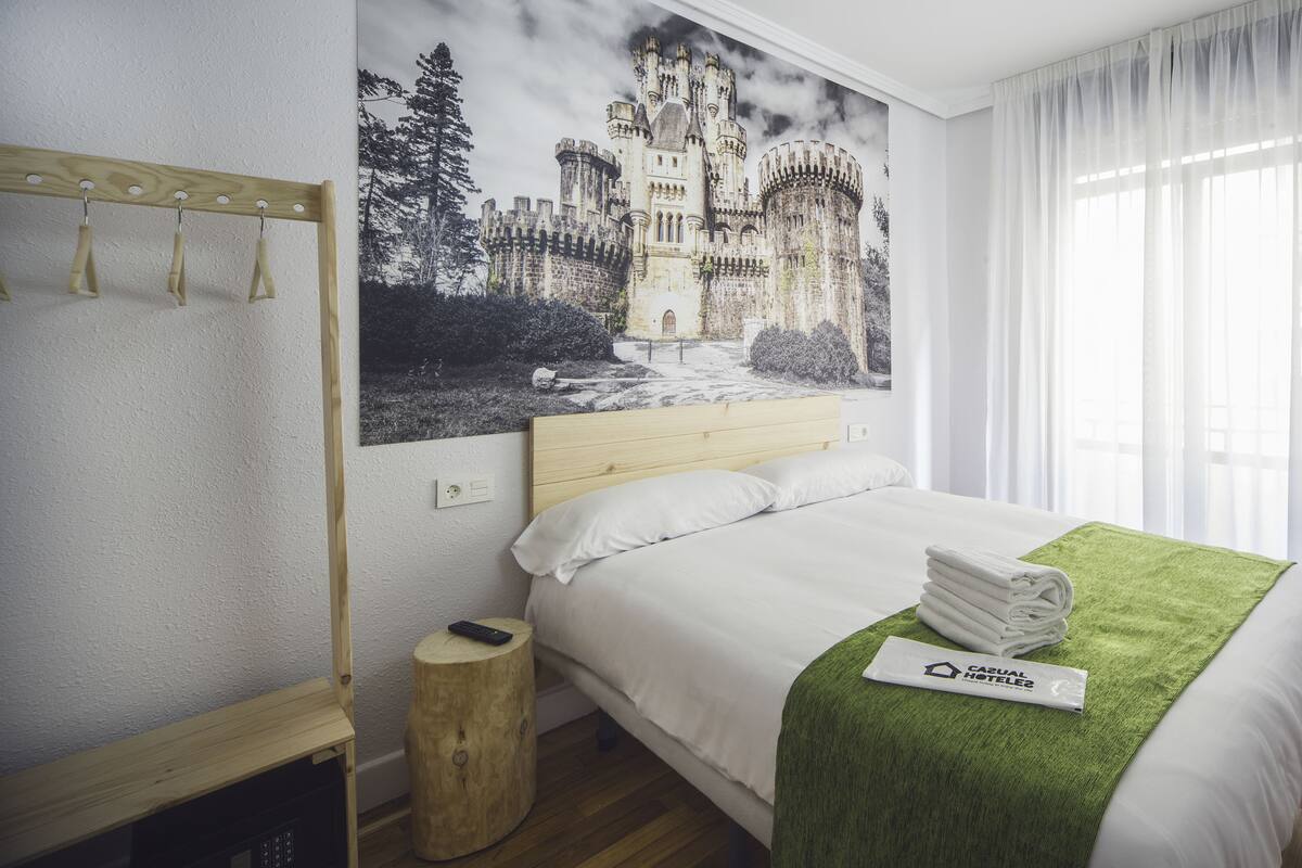 Habitación de hotel con una cama, una mesita de noche de madera, un perchero para toallas en la pared, un control remoto y un cuadro con temática de castillo.