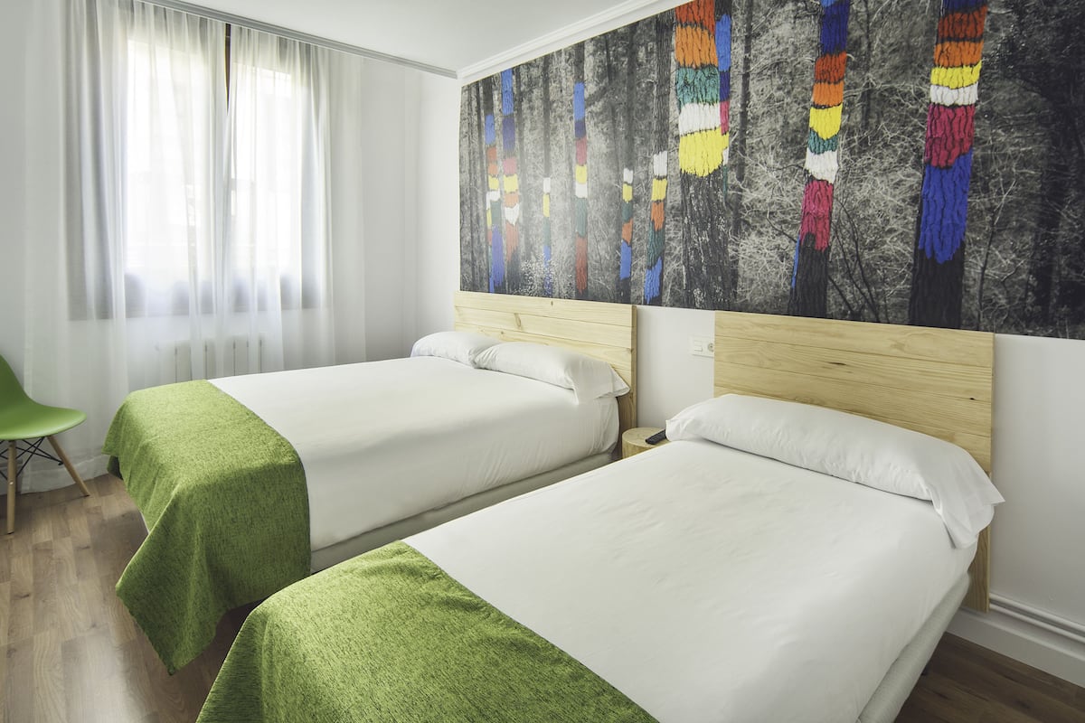 Habitación de hotel con dos camas, una colcha verde, un cuadro abstracto grande en la pared y una ventana con cortinas translúcidas.