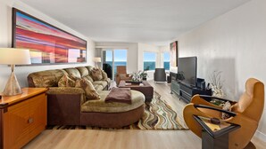 A modern living room with a large sofa, a coffee table, and a TV. There is a view of the ocean through large windows.