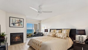 A modern bedroom with a large bed, a fireplace, a ceiling fan, and a view of the ocean.