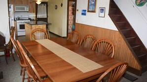 A dining area with a wooden table and chairs, a kitchen in the background, and a staircase.
