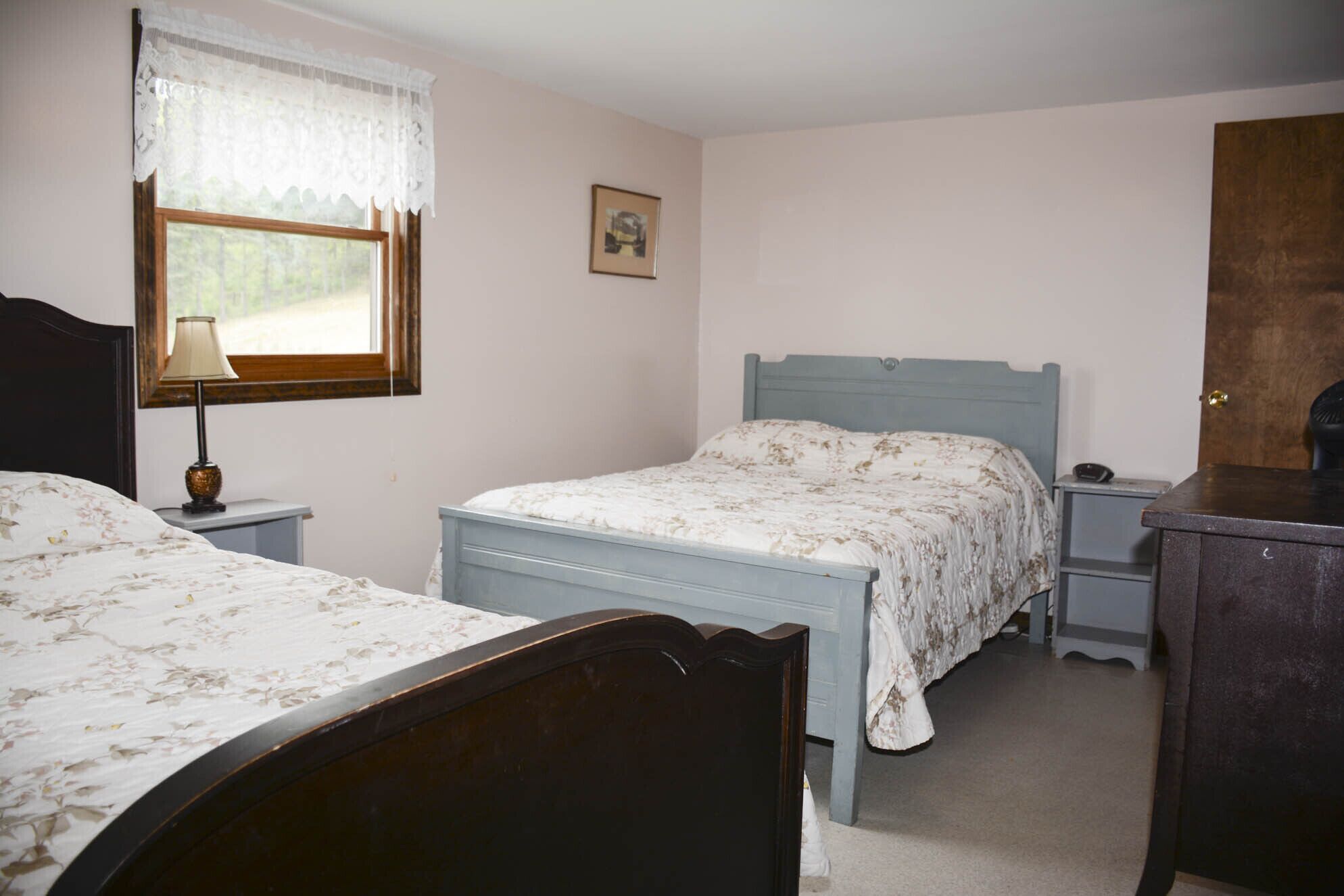 A bedroom with two single beds, a nightstand with a lamp, a dresser, and a window with lace curtains.