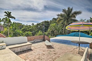 A rooftop pool area with a colorful umbrella, lounge chairs, and a view of lush greenery.