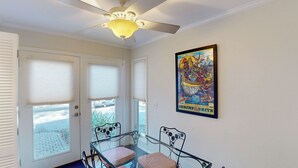 Ein Zimmer mit Deckenventilator, einem Glastisch mit zwei Stühlen, einer Tür mit Jalousien und einem gerahmten Poster an der Wand.