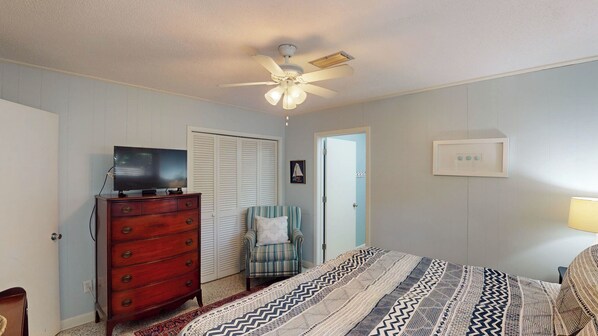 A bedroom with a ceiling fan, a television on a dresser, a chair, a bed with patterned bedding, and a door with a light switch.
