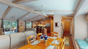 A spacious living area with a wooden dining table, comfortable seating, a kitchen in the background, and a ceiling fan.