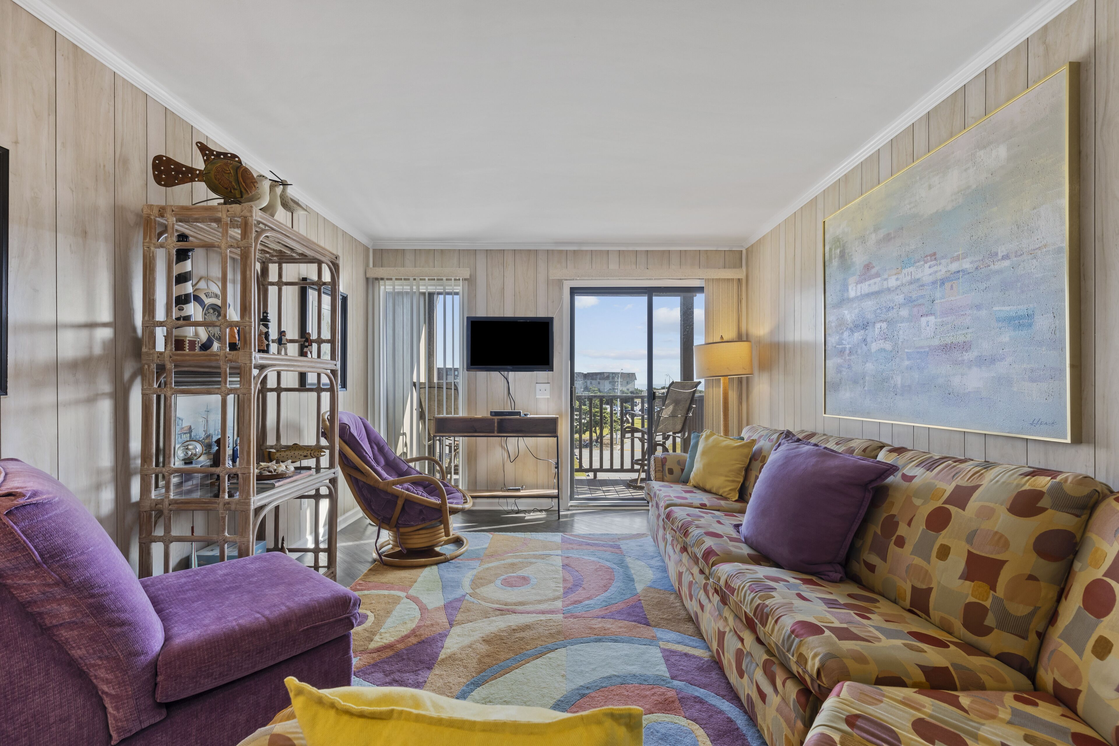 A living room with a purple sofa, a patterned rug, a wooden bookshelf, and a large painting on the wall.