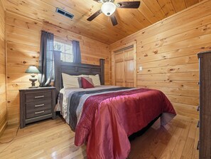 A wooden cabin bedroom with a bed, nightstands, a ceiling fan, and a window with curtains.