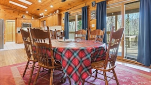 A wooden cabin interior with a dining table set for four, wooden chairs, a kitchen area, and large windows with curtains.