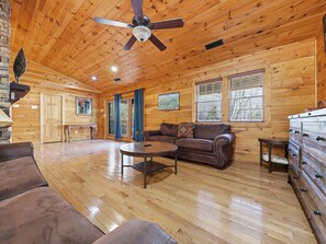 A wooden cabin interior with a wooden ceiling, a ceiling fan, a brown leather sofa, a coffee table, and a dresser with drawers.