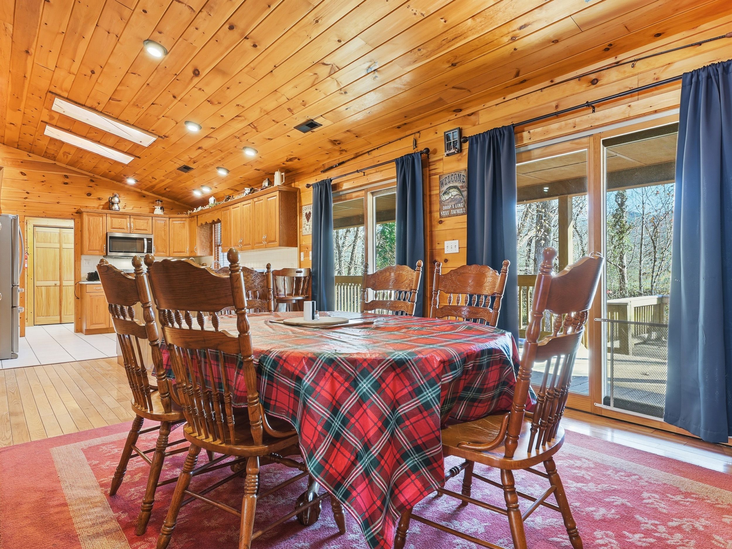 A wooden cabin interior with a dining table set for four, wooden chairs, a kitchen area, and large windows with curtains.