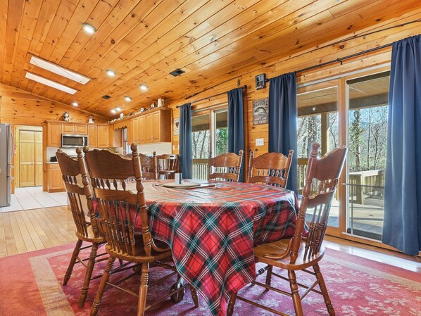 A wooden cabin interior with a dining table set for four, wooden chairs, a kitchen area, and large windows with curtains.
