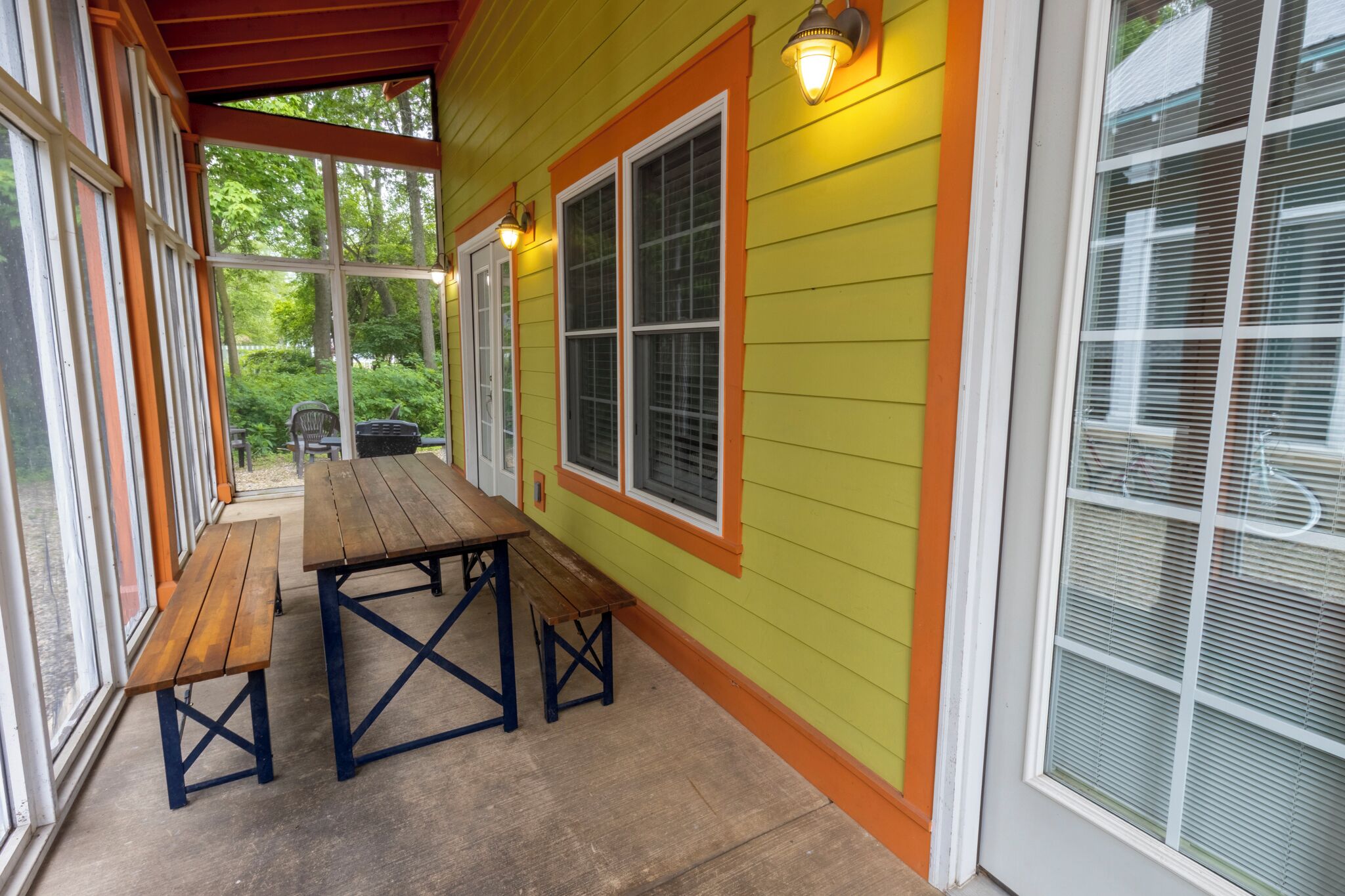 A covered porch with a wooden table and benches, large windows, and a yellow exterior wall.
