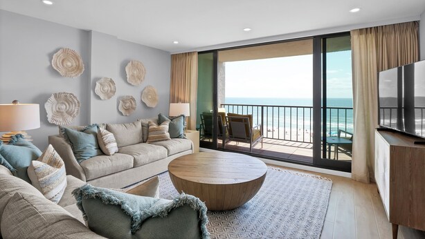 Een moderne woonkamer met een grote bank, een ronde salontafel en een televisie. Er is een balkon met uitzicht op de oceaan.