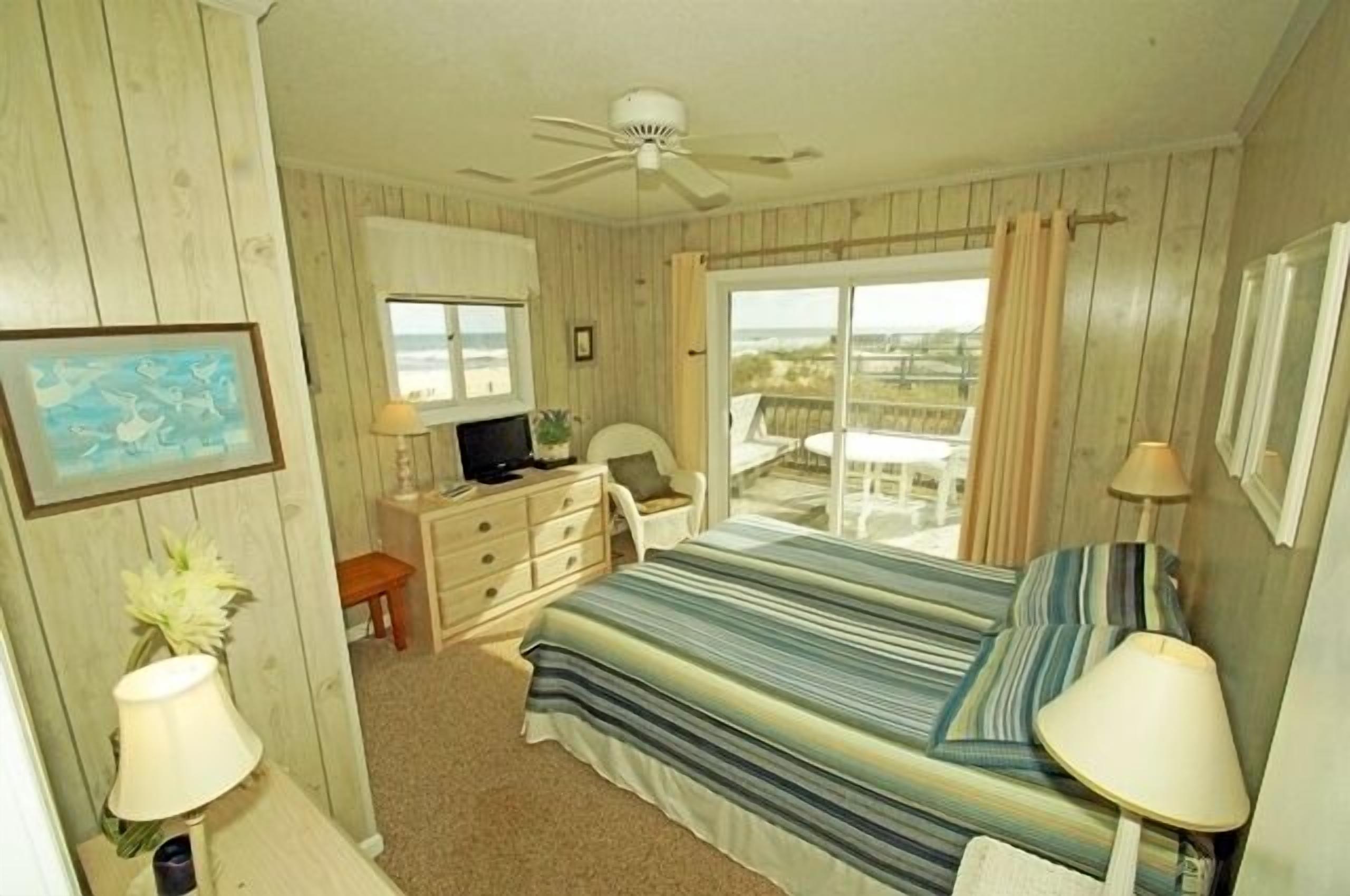 Une chambre à coucher comprenant un lit, un bureau, une chaise, un ventilateur de plafond et une fenêtre donnant sur la plage.